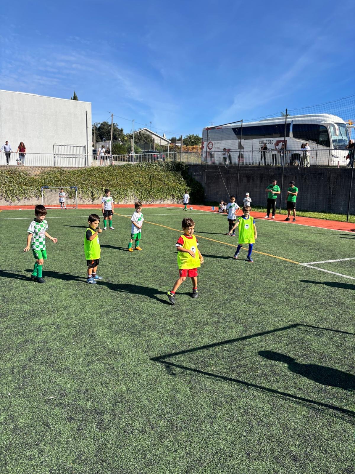 Crianças da Escola de Futebol em campo, com os pais a assistir ao fundo