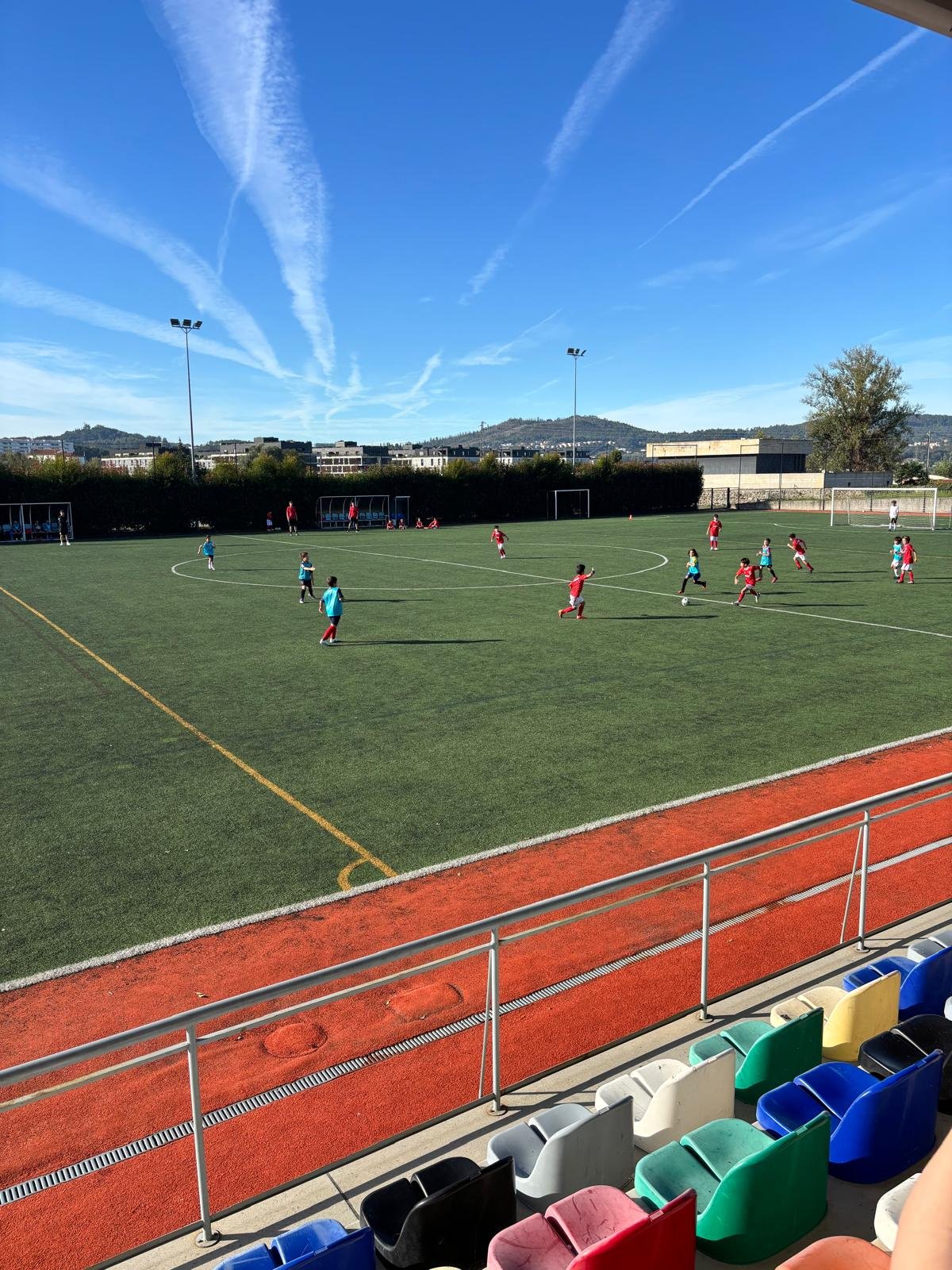 Campo de futebol visto da bancada, com crianças a jogar numa manhã de céu azul