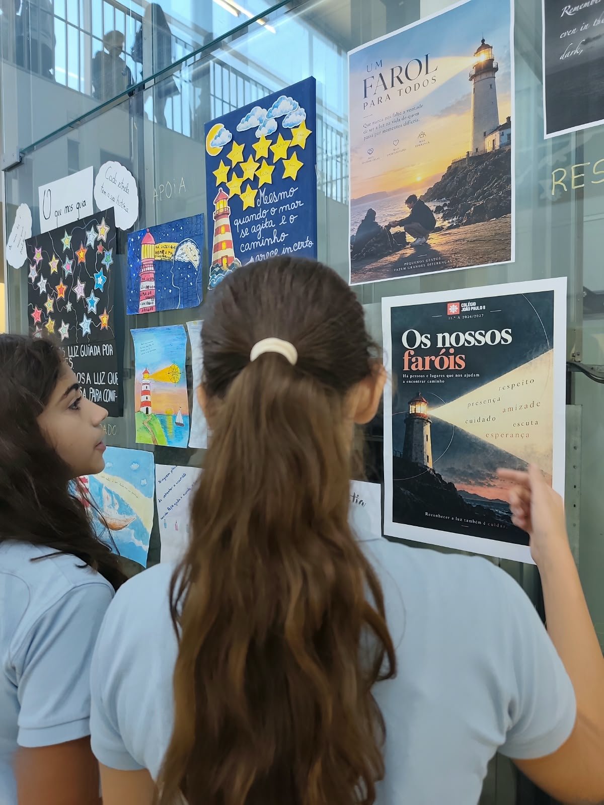 Duas alunas a ler o cartaz «Os nossos faróis», num vidro com trabalhos expostos
